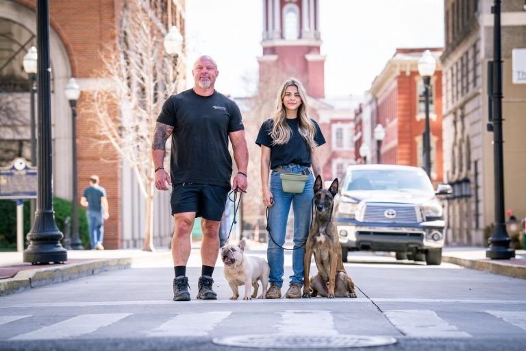 Two people walking dogs on a city street with brick buildings in the background.