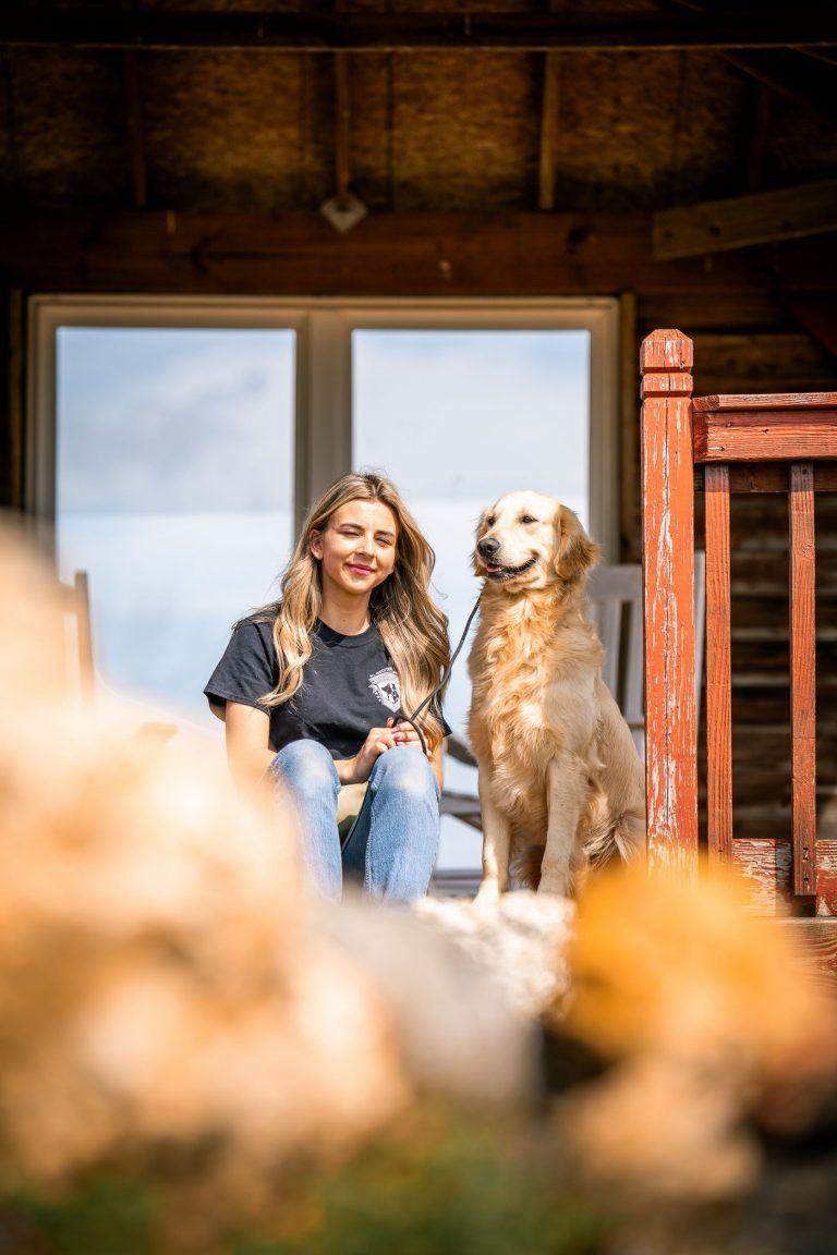 Woman sitting with a golden retriever in a rustic doorway, with a blurred foreground of flowers.
