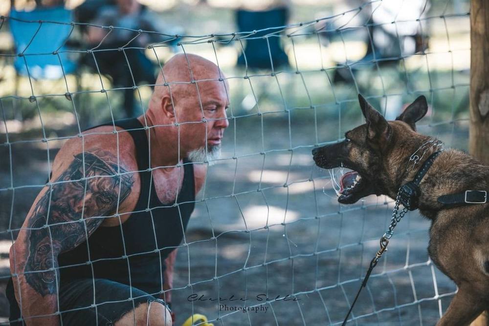 Man crouches behind a net facing a barking dog on a leash in an outdoor training area