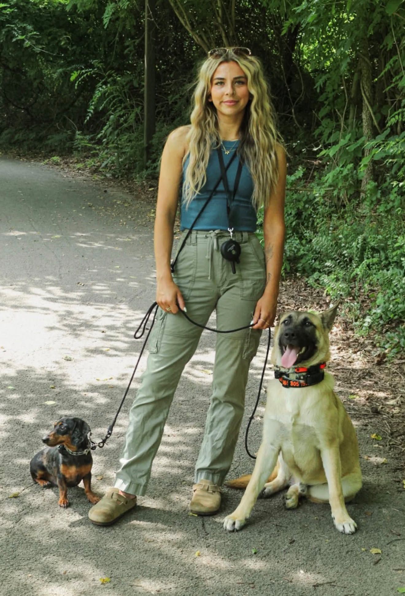 Rebeka, dog trainer at K9 Services Unlimited, standing on a shaded path with a dachshund and a large tan dog on leashes